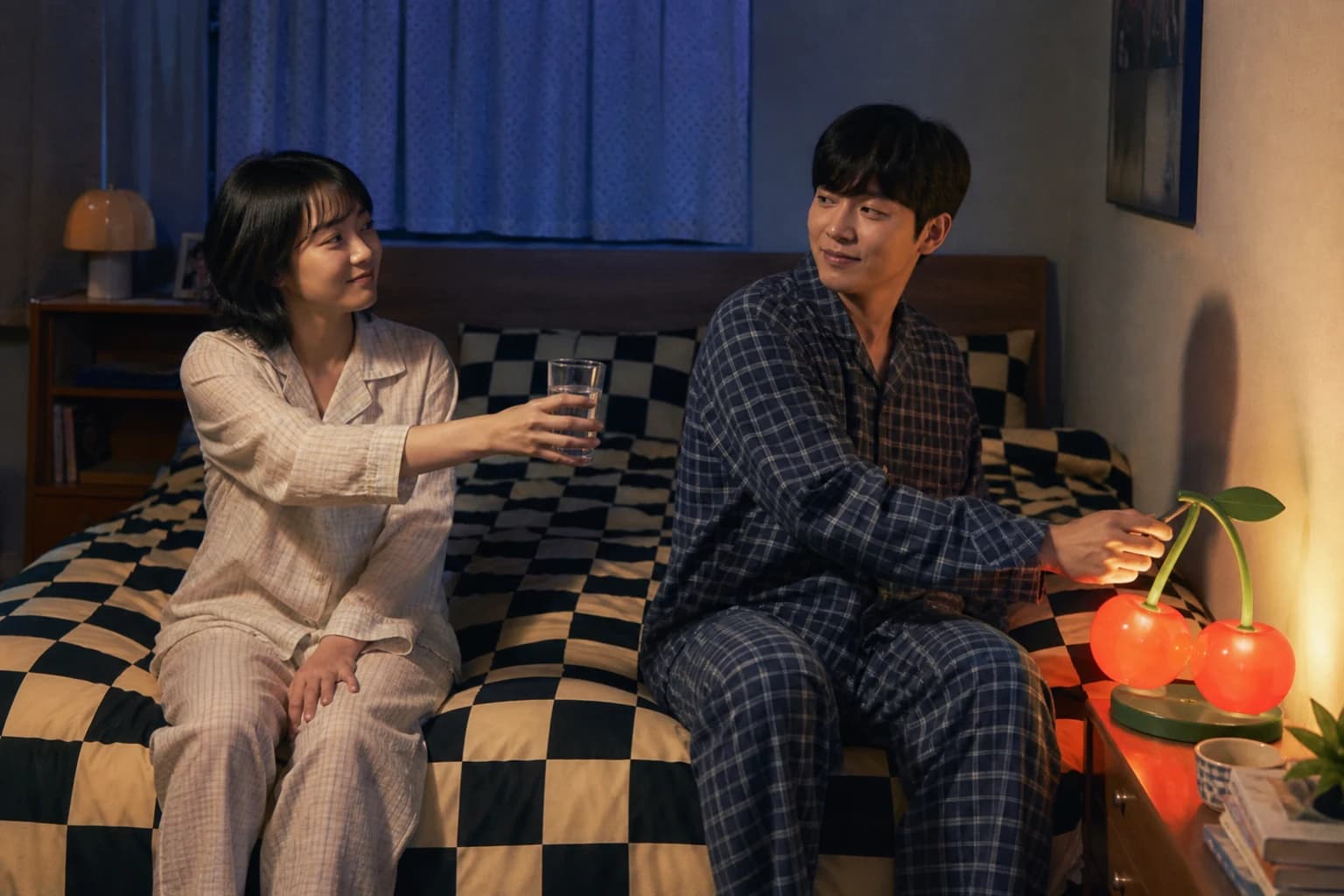 체커보드 이불 끝에 앉아 물잔을 건네며 어색하게 웃는 성인 커플